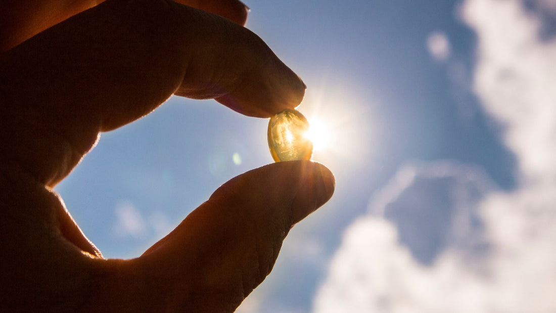 Hand holding a vitamin D capsule up to the sunlight with a bright sky in the background.